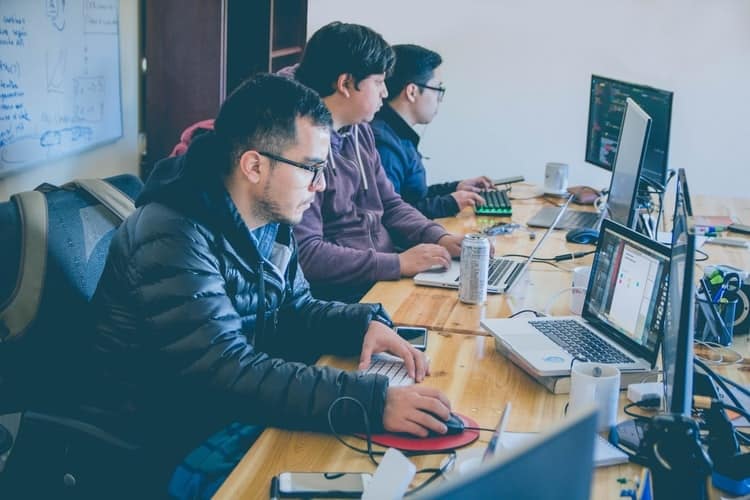 tres homens sentados em frente a computadores com codigos de programacao na tela