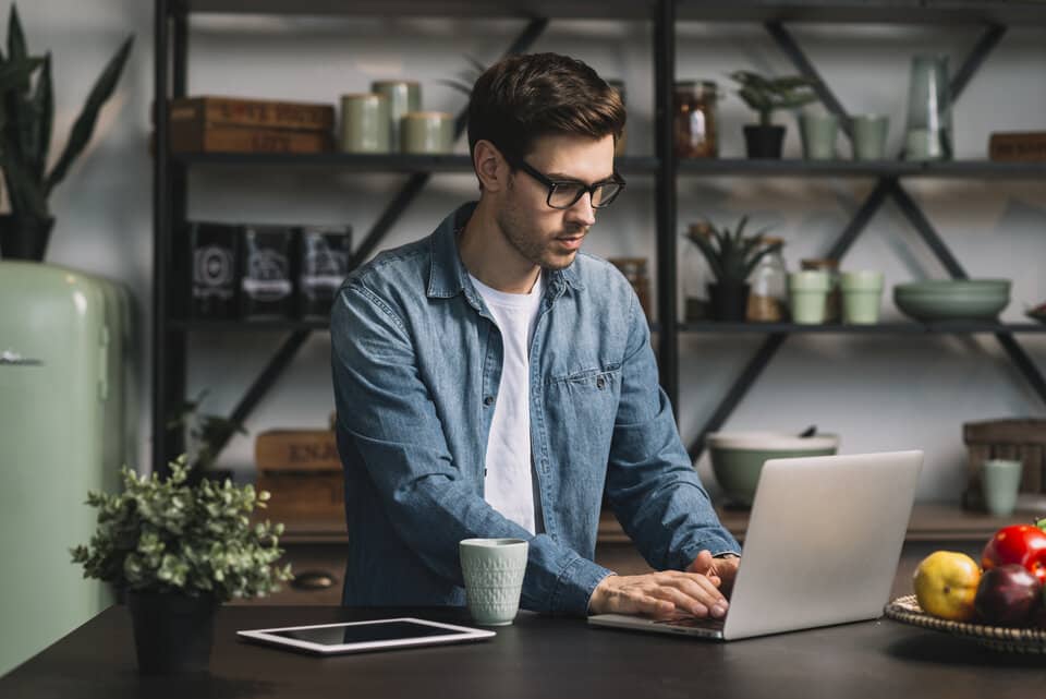 Homem de óculos usando notebook na mesa com uma cozinha ao fundo