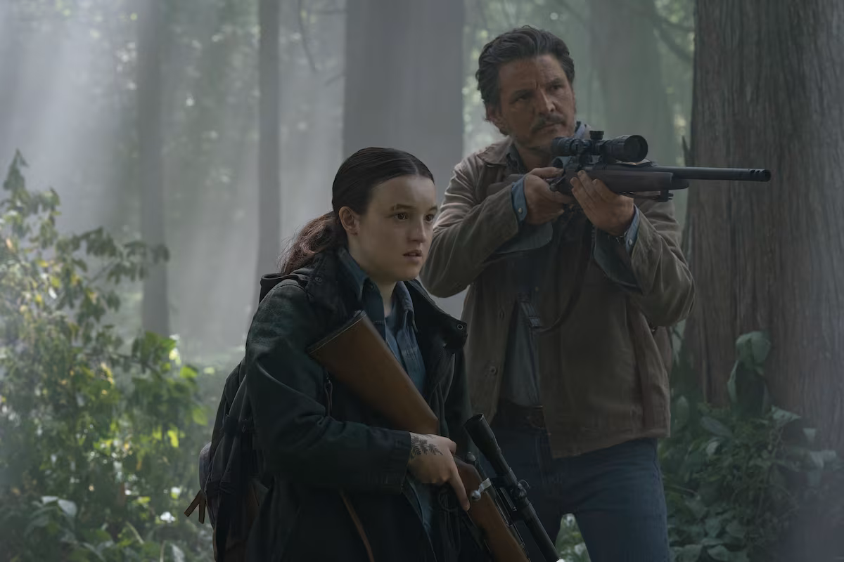 Ellie e Joel, de The Last of Us, caminham por uma floresta coberta de vegetação. Ellie carrega uma arma, enquanto Joel observa a área com um rifle.