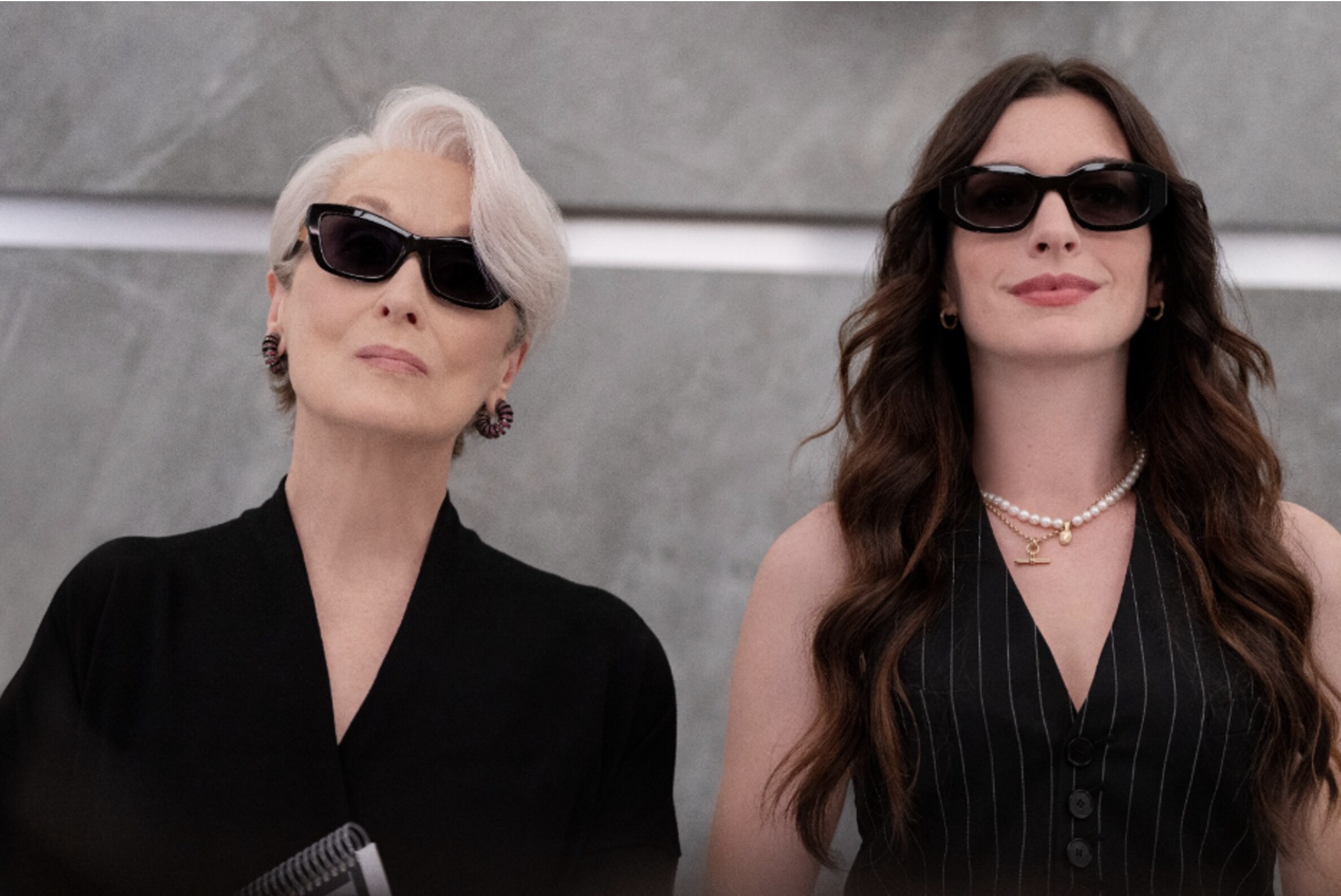 Cena do filme ''O Diabo Veste Prada 2'' com duas mulheres usando roupas formais e óculos escuros, lado a lado, em um ambiente corporativo.